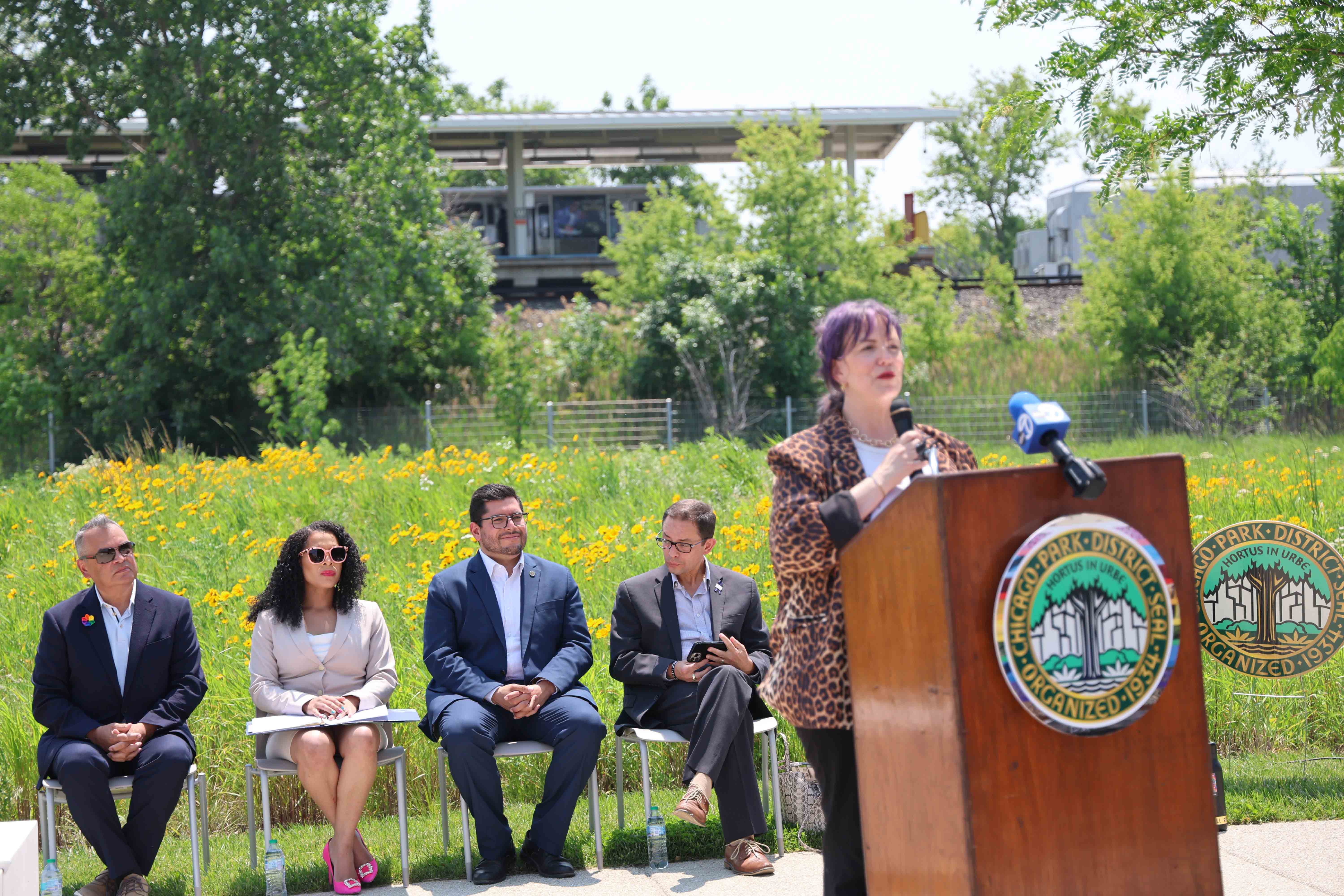 A woman speaks at a podium, with four seated people behind her and a train station in the background.